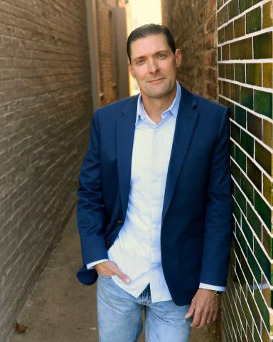 Image of Charles Donlea, leaning against a brick wall wearing a blue sports coat, white shirt, and jeans.
